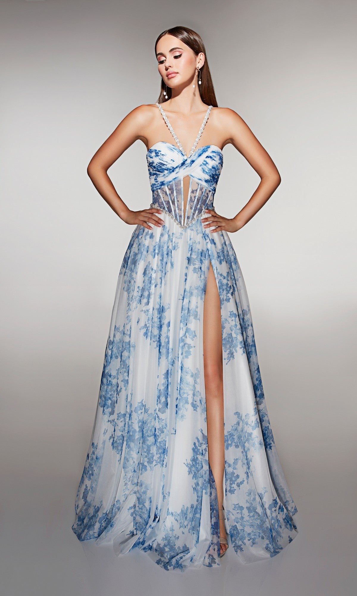 Woman in the Alyce 61994 long prom dress, featuring blue and white floral print, a sheer corset bodice, crisscross straps, and high slit, stands with hands on hips against a plain background.